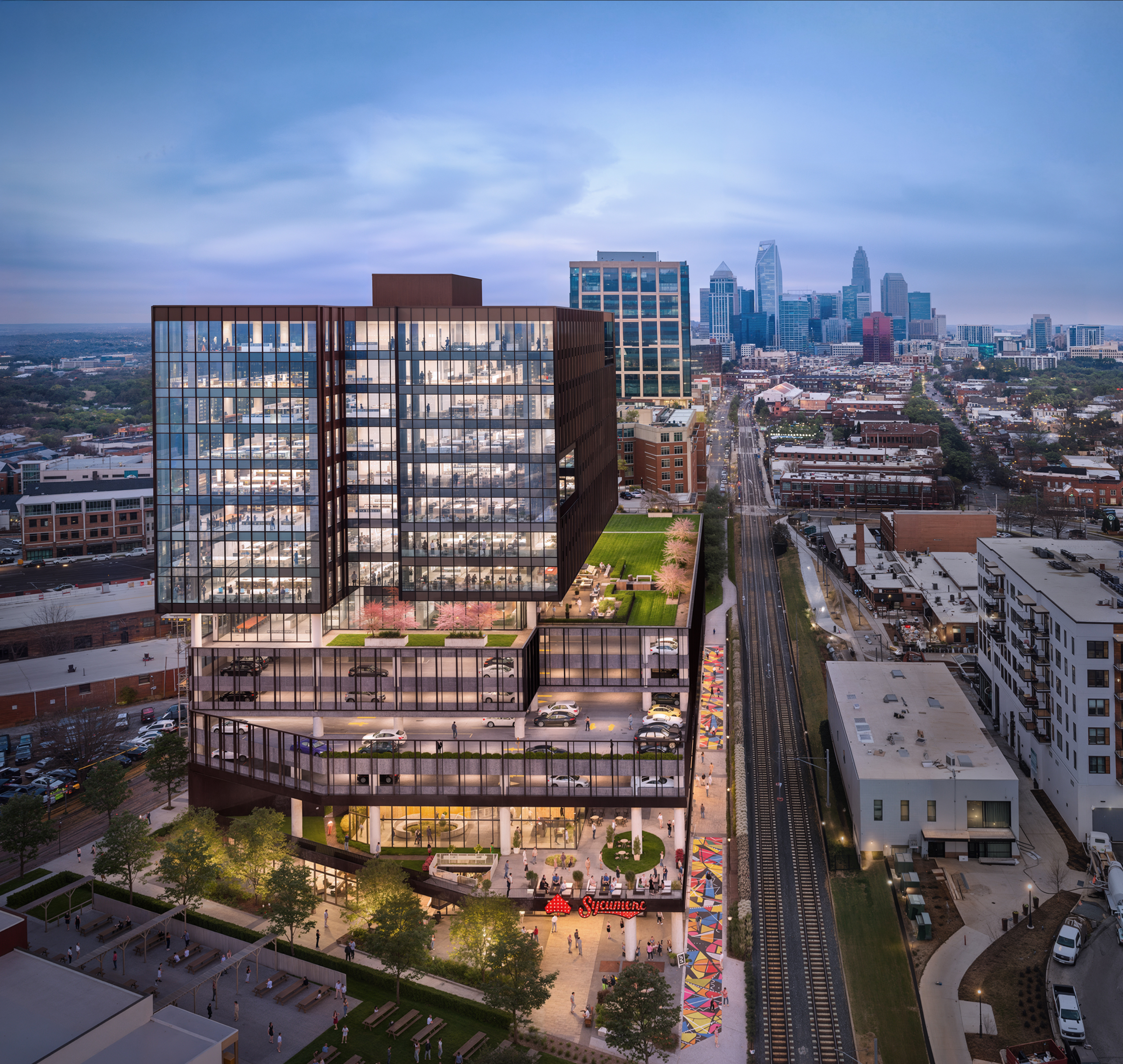 The Line 2151 Hawkins Charlotte mixed-use development aerial rendering by Bogza.com. A twilight drone view of the 16-story Class A office building in South End, featuring a modern glass tower above a massive parking podium, a green rooftop terrace, ground-floor Sycamore Brewing, the adjacent Rail Trail, and the Uptown Charlotte skyline in the background.