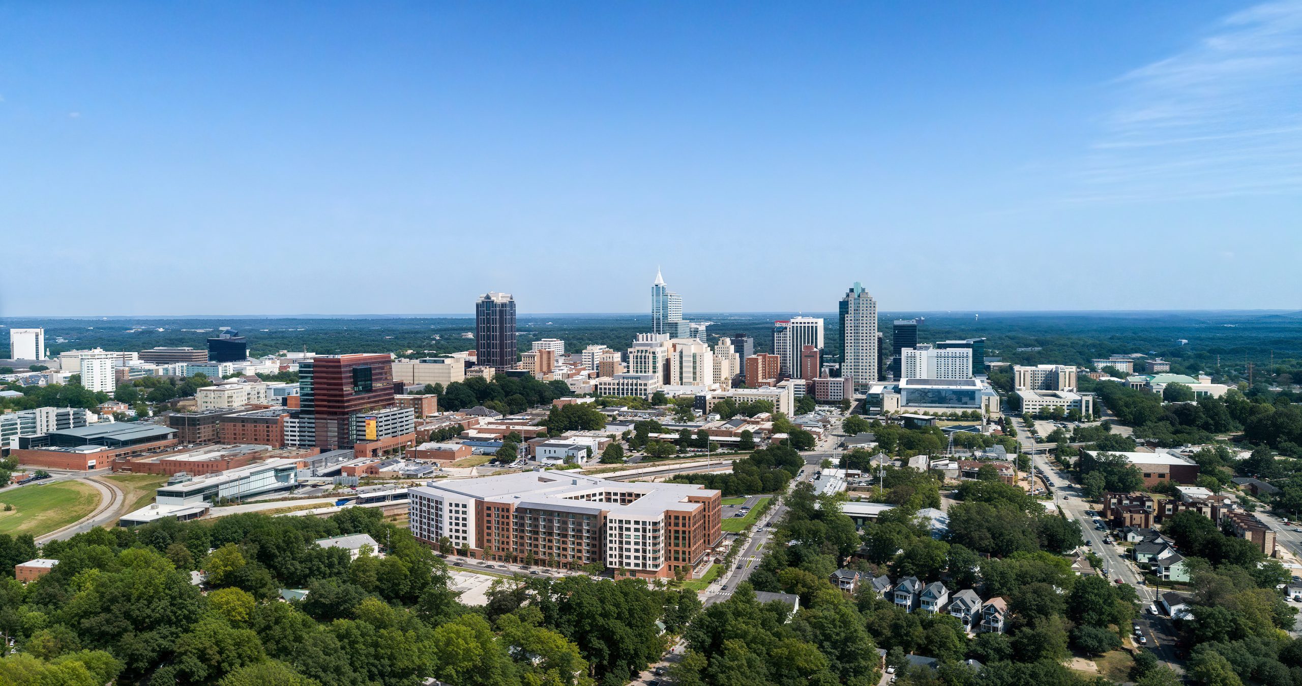 Platform Raleigh West End mixed-use development aerial architectural rendering by Bogza.com. A high-angle daytime bird's-eye view of the downtown Raleigh skyline, showcasing the newly redeveloped Clancy & Theys headquarters site, featuring the expansive brick and white Platform apartment building with its central courtyard and integrated parking deck.