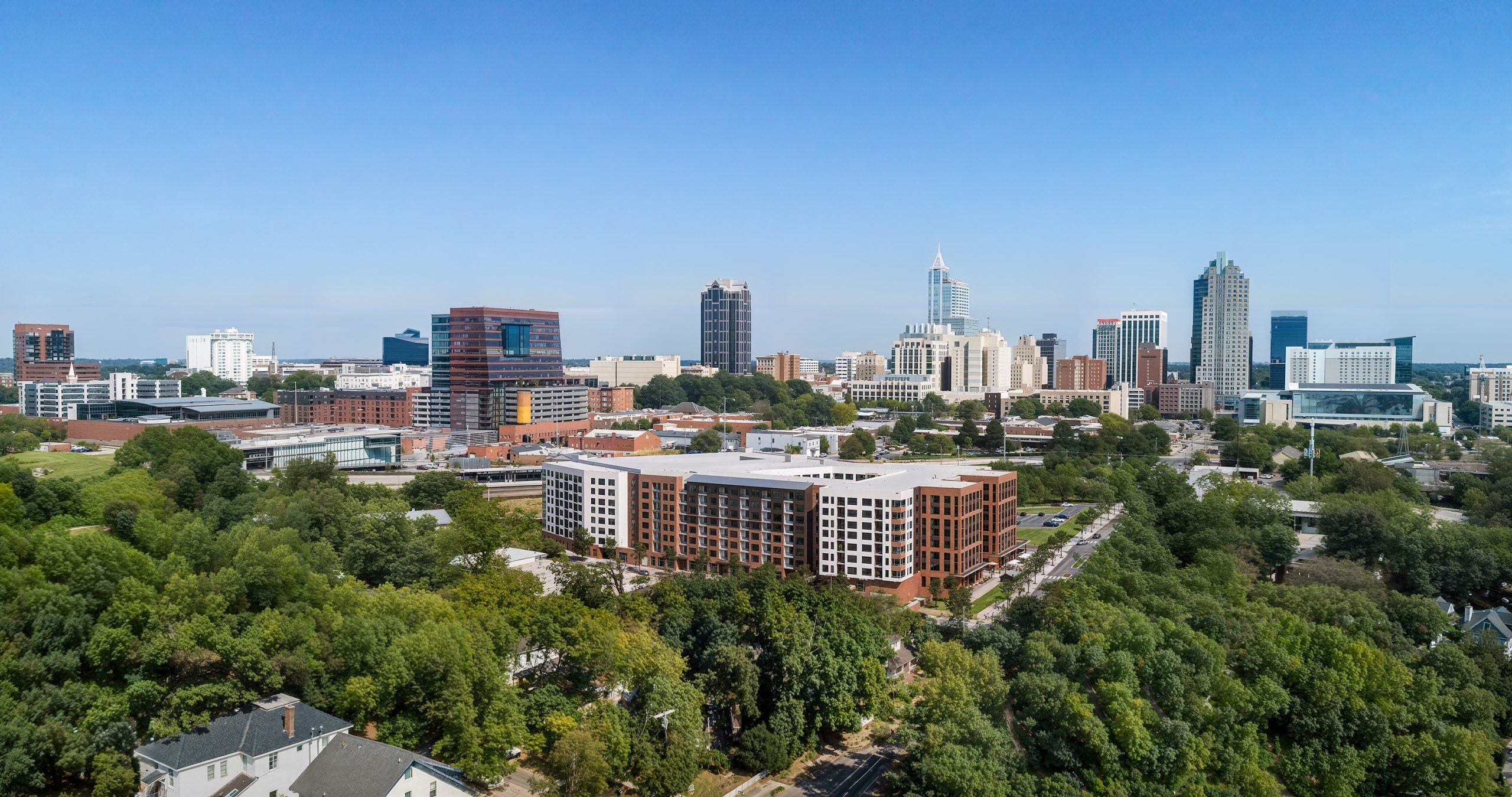 Platform apartments Raleigh NC downtown skyline aerial rendering by Bogza.com. A stunning daytime drone view of the 7-story mixed-use residential community in the West End neighborhood, highlighting the building's U-shaped design, ground-floor retail spaces, and its proximity to the towering downtown Raleigh skyline.