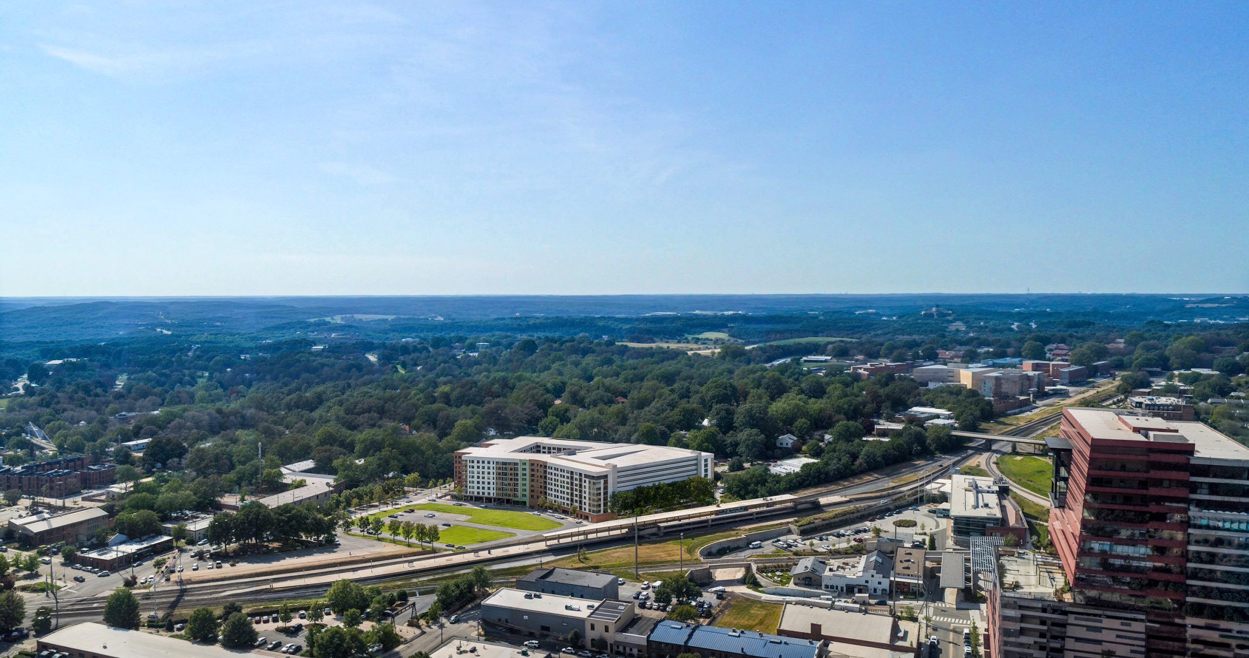 Platform Raleigh Union Station mixed-use development aerial rendering by Bogza.com. A wide daytime aerial perspective looking over the Platform apartment community, showcasing its location directly adjacent to the railroad tracks and Raleigh Union Station, surrounded by lush green tree canopies.