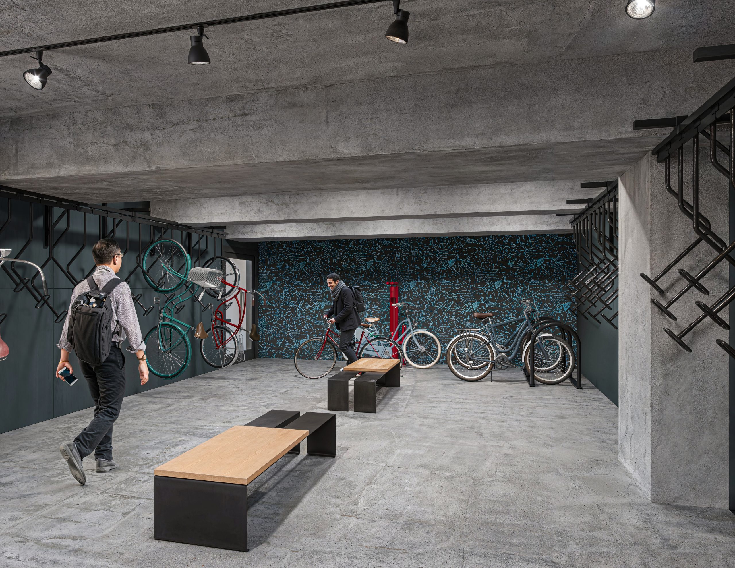 Tower 3 at Bloc 83 Raleigh office bike room interior rendering by Bogza.com. A modern, industrial-style tenant bicycle storage facility featuring exposed concrete ceilings and walls, wall-mounted vertical bike racks, sleek wooden benches, and a vibrant blue patterned accent wall.