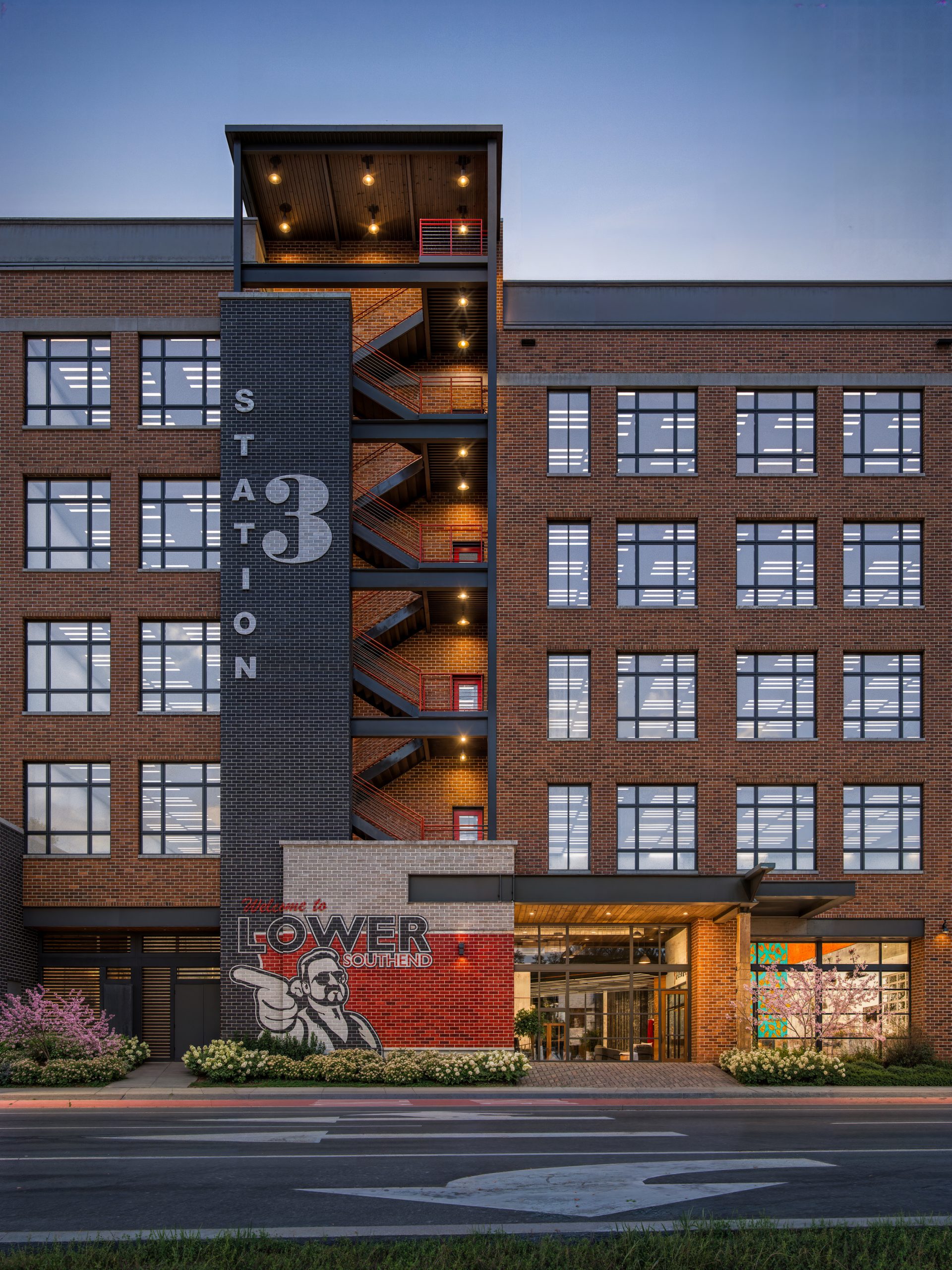 The Station at LoSo Charlotte office architecture exterior rendering by Bogza.com. A striking twilight close-up of Station 3's exterior, highlighting the red brick warehouse-style facade, large grid windows, and the prominent black steel exterior staircase column.