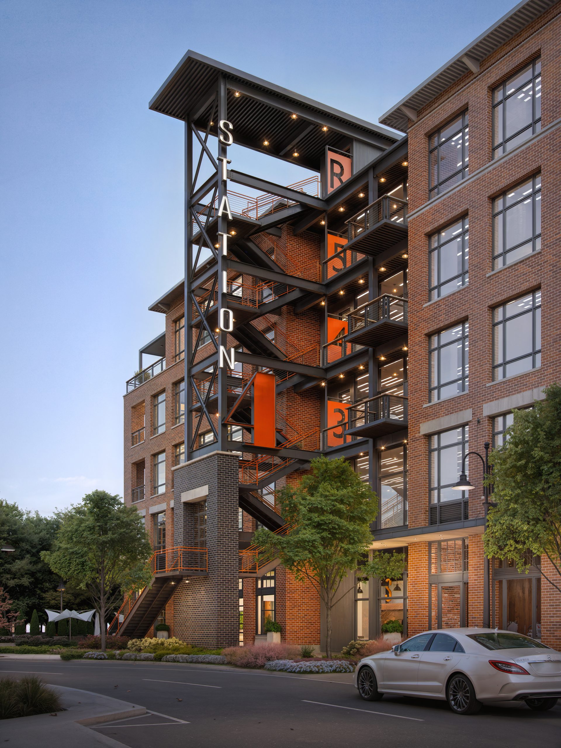Station 4 at The Station at LoSo Charlotte exterior architectural rendering by Bogza.com. A twilight street-level view of the modern 5-story creative office building, showcasing a distinctive black steel exterior staircase structure with bright orange landing accents and a red brick facade.