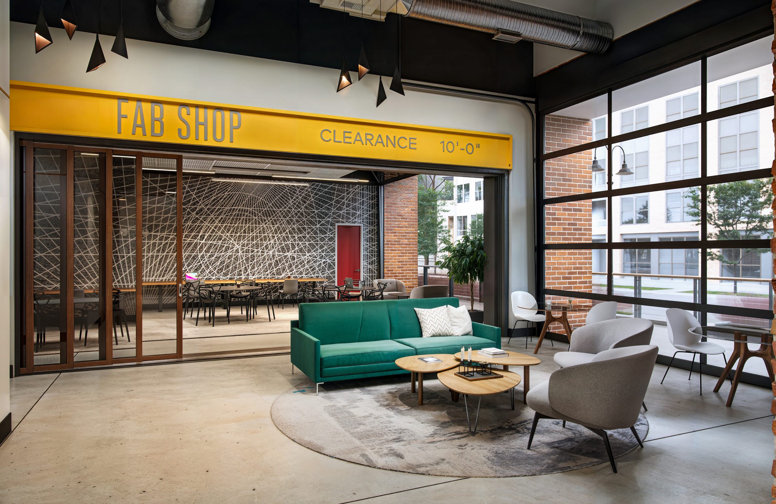 Station 3 at The Station at LoSo Charlotte FAB SHOP lounge interior rendering by Bogza.com. A contemporary indoor-outdoor amenity space featuring a green sofa, wood-paneled walls, an open garage door leading to an exterior courtyard, and modern industrial lighting.