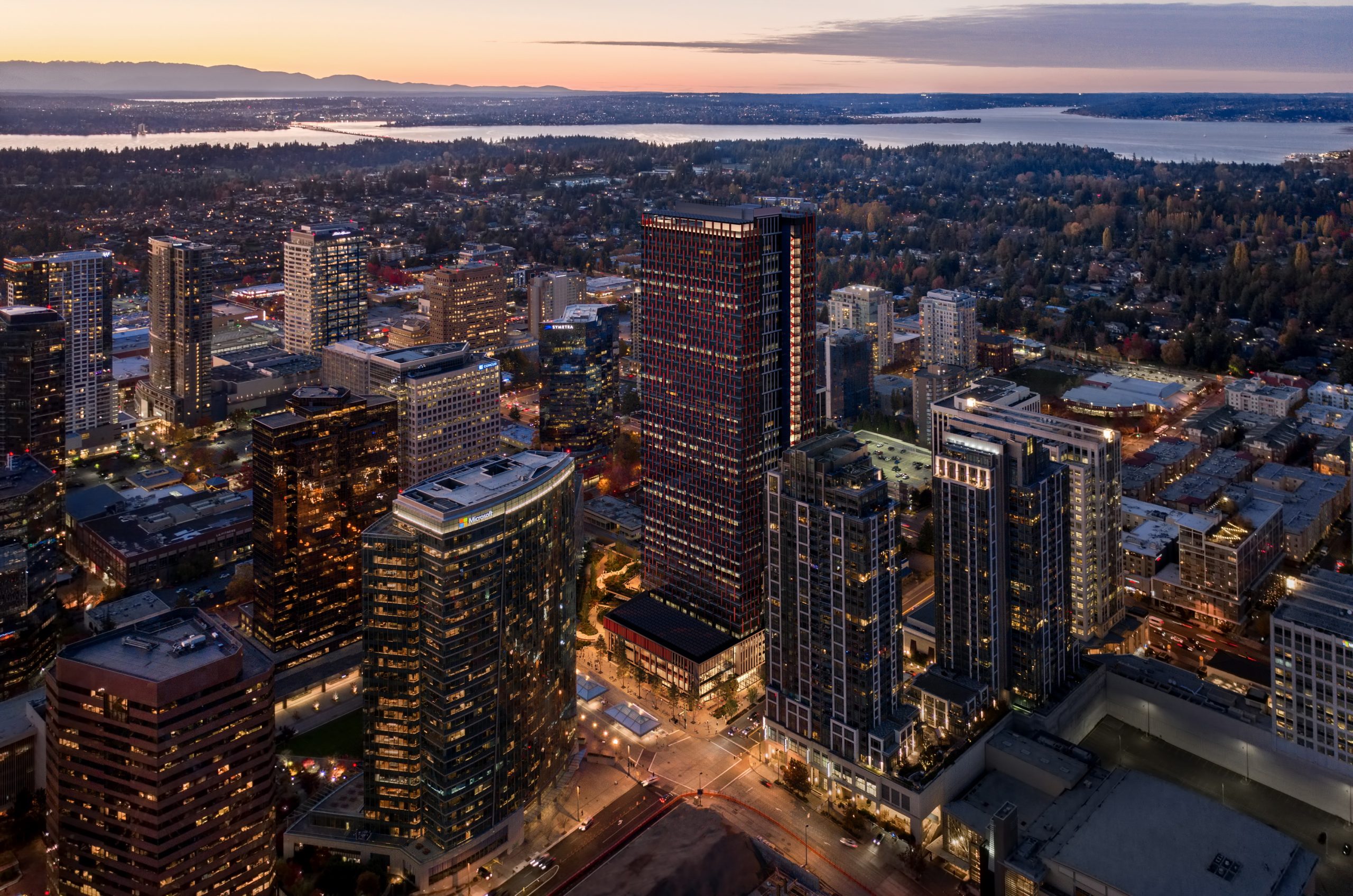 600 Bellevue twilight aerial rendering by Bogza.com. A high-angle drone perspective of the modern 31-story Amazon office tower standing prominently in the illuminated downtown Bellevue skyline with waterfront views.