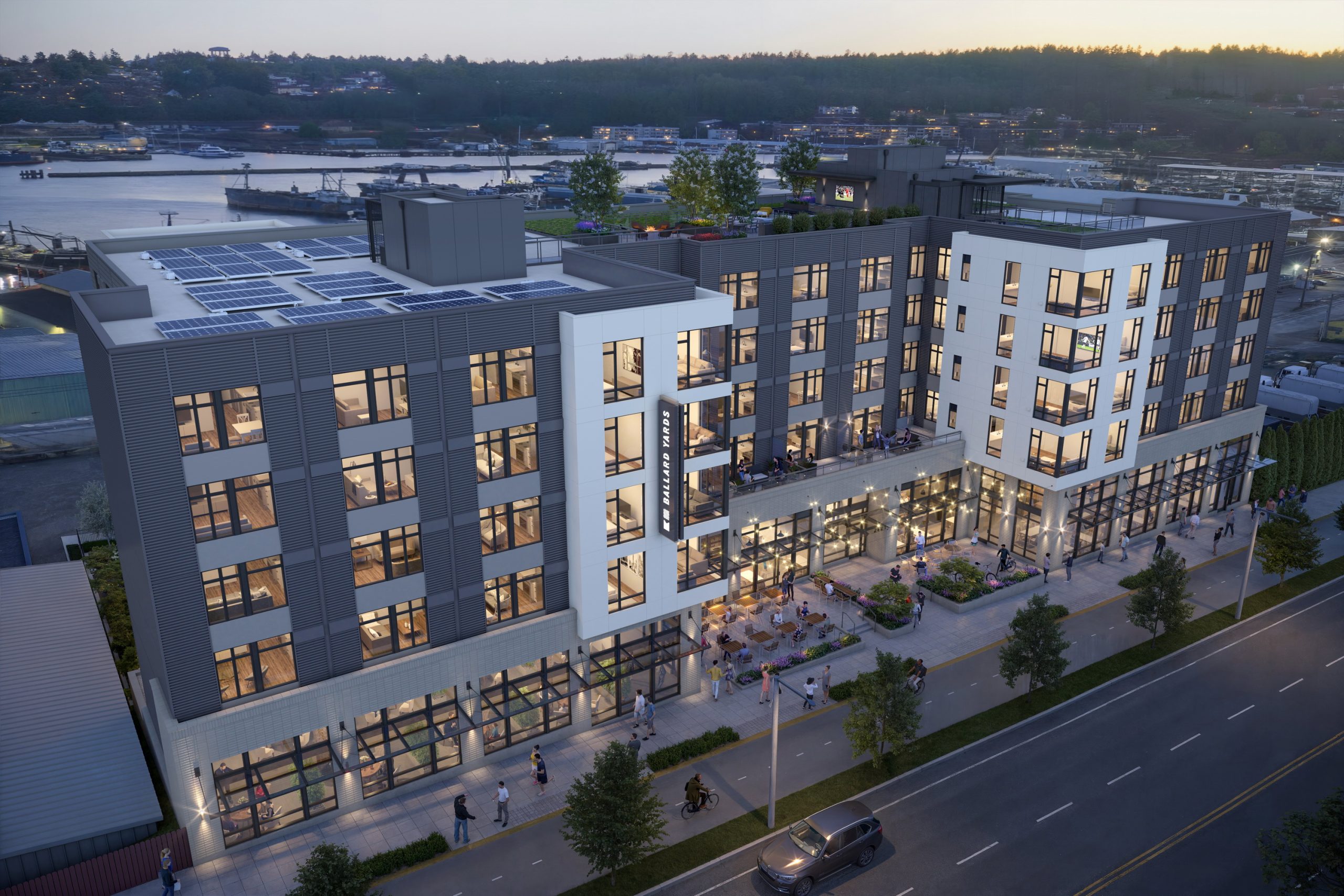 Ballard Yards Seattle aerial architectural exterior rendering by Bogza.com. A dusk view of the 7-story mixed-use apartment building on Market Street, featuring a modern facade, rooftop deck, solar panels, and an active pedestrian streetscape.