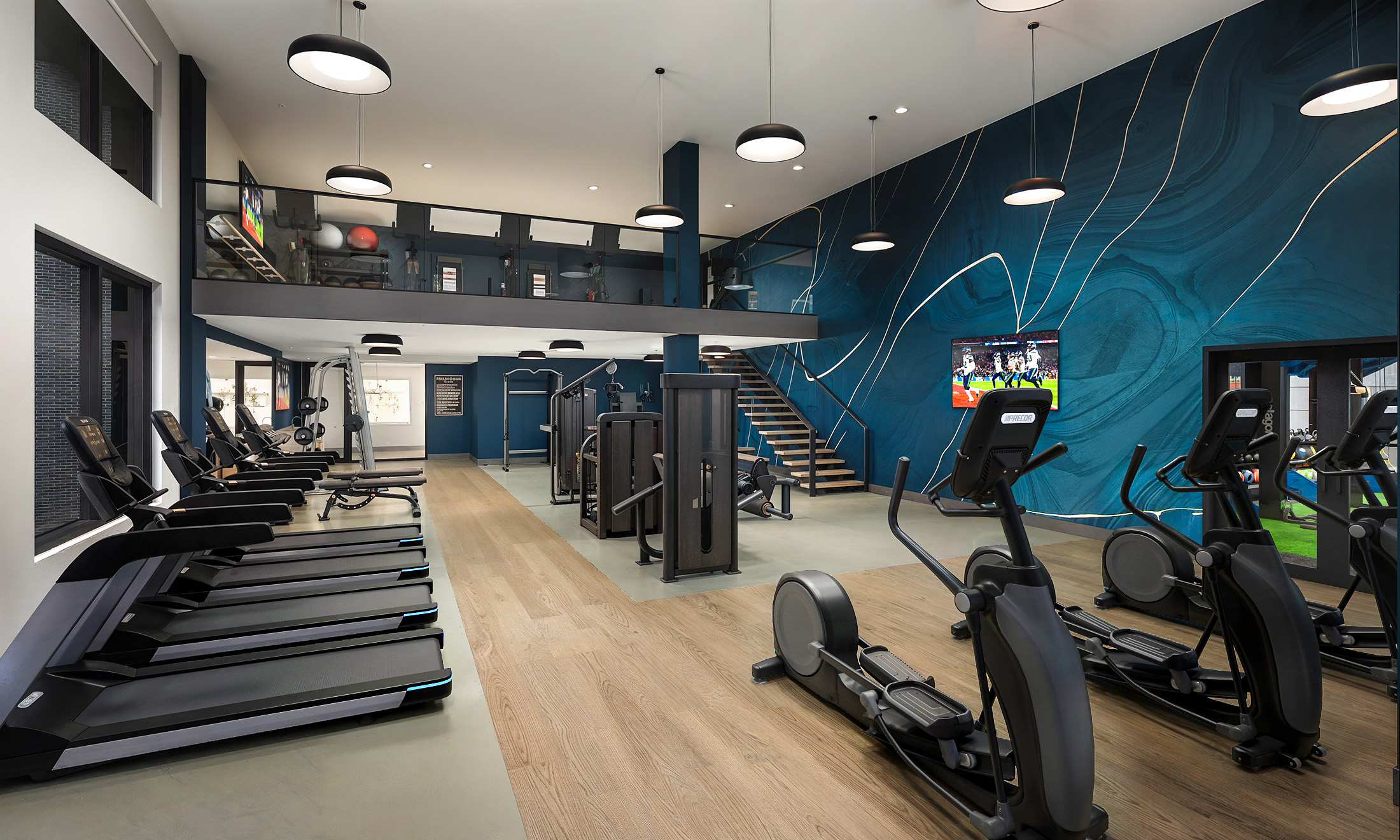 Broadstone Strata Seattle fitness center interior rendering by Bogza.com. A spacious, multi-level gym amenity featuring state-of-the-art cardio treadmills and ellipticals, weight machines, a staircase to the upper level, and a striking blue graphic accent wall.