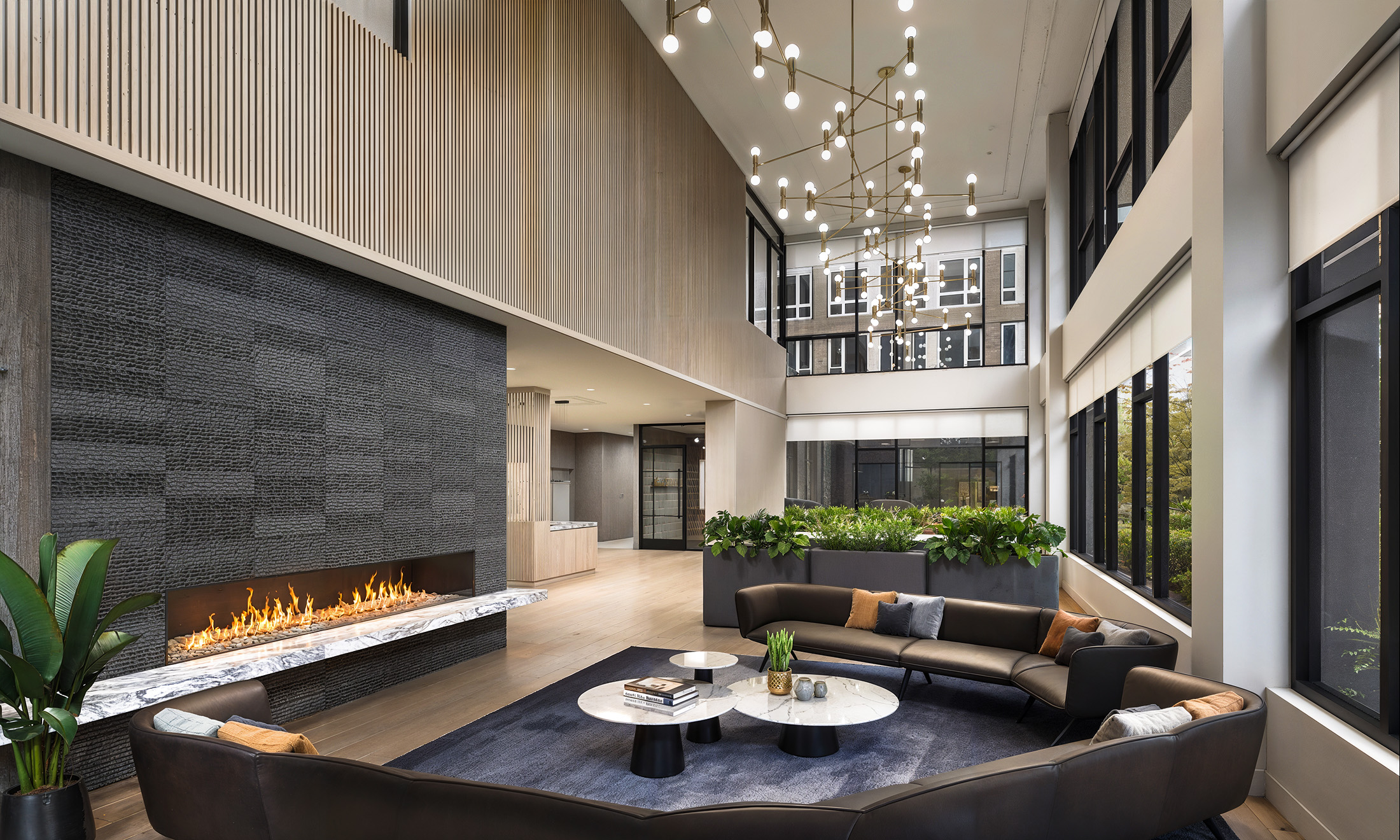 Broadstone Strata Seattle luxury lobby interior architectural rendering by Bogza.com. A stunning double-height community lounge featuring a massive dark textured stone fireplace, a modern brass chandelier, a large curved leather sofa, vertical wood slat walls, and large windows.