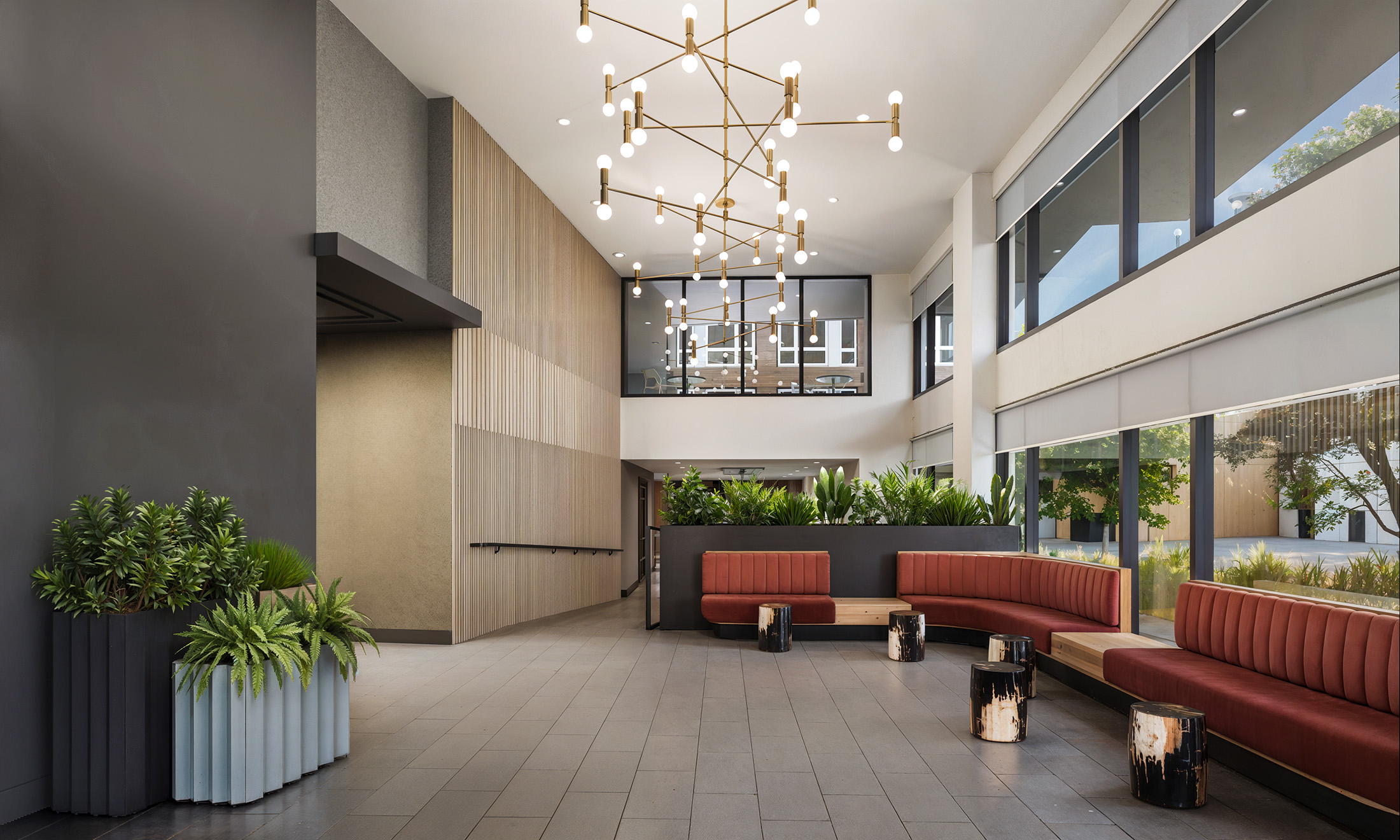 Broadstone Strata Seattle modern concierge lobby interior rendering by Bogza.com. A bright and inviting reception area featuring a long, curved red upholstered bench, natural wood accent tables, modern lighting, lush planter boxes, and expansive street-facing windows.