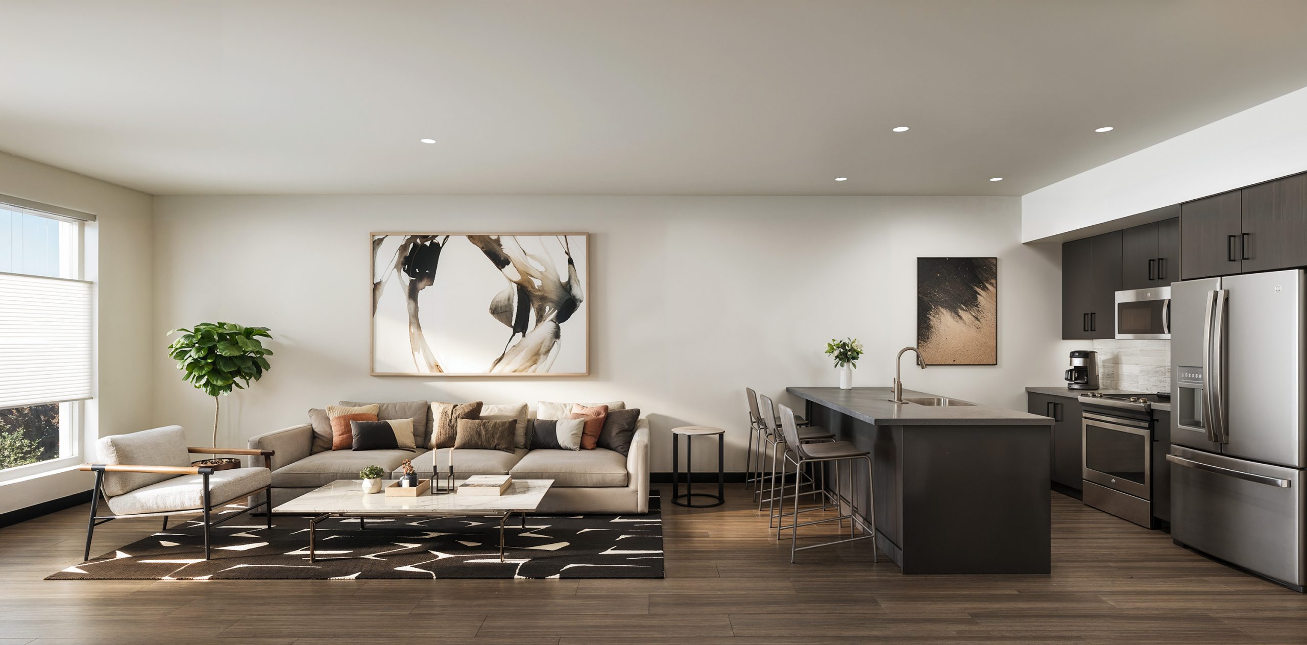 Broadstone Strata Seattle luxury apartment interior rendering by Bogza.com. A bright, modern open-concept living room and kitchen in the Ravenna neighborhood, featuring wood-style flooring, a cozy lounge area with contemporary artwork, and a sleek kitchen with dark cabinetry, quartz countertops, and stainless steel appliances.