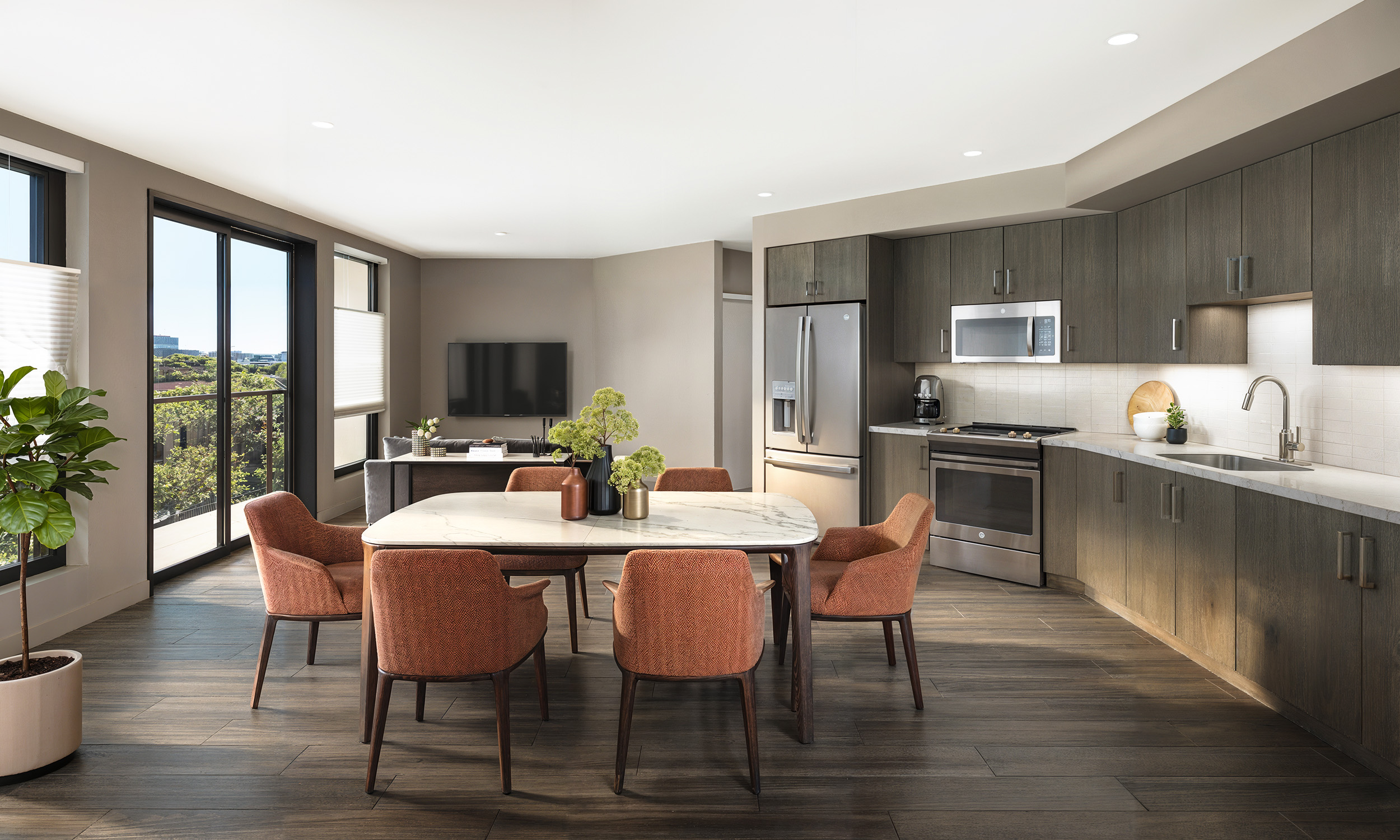 Broadstone Strata Seattle modern apartment dining and kitchen interior architectural rendering by Bogza.com. A spacious open floor plan featuring a marble dining table with orange accent chairs, wood-style flooring, and a contemporary kitchen showcasing flat-panel wood cabinetry, quartz countertops, and stainless steel appliances.
