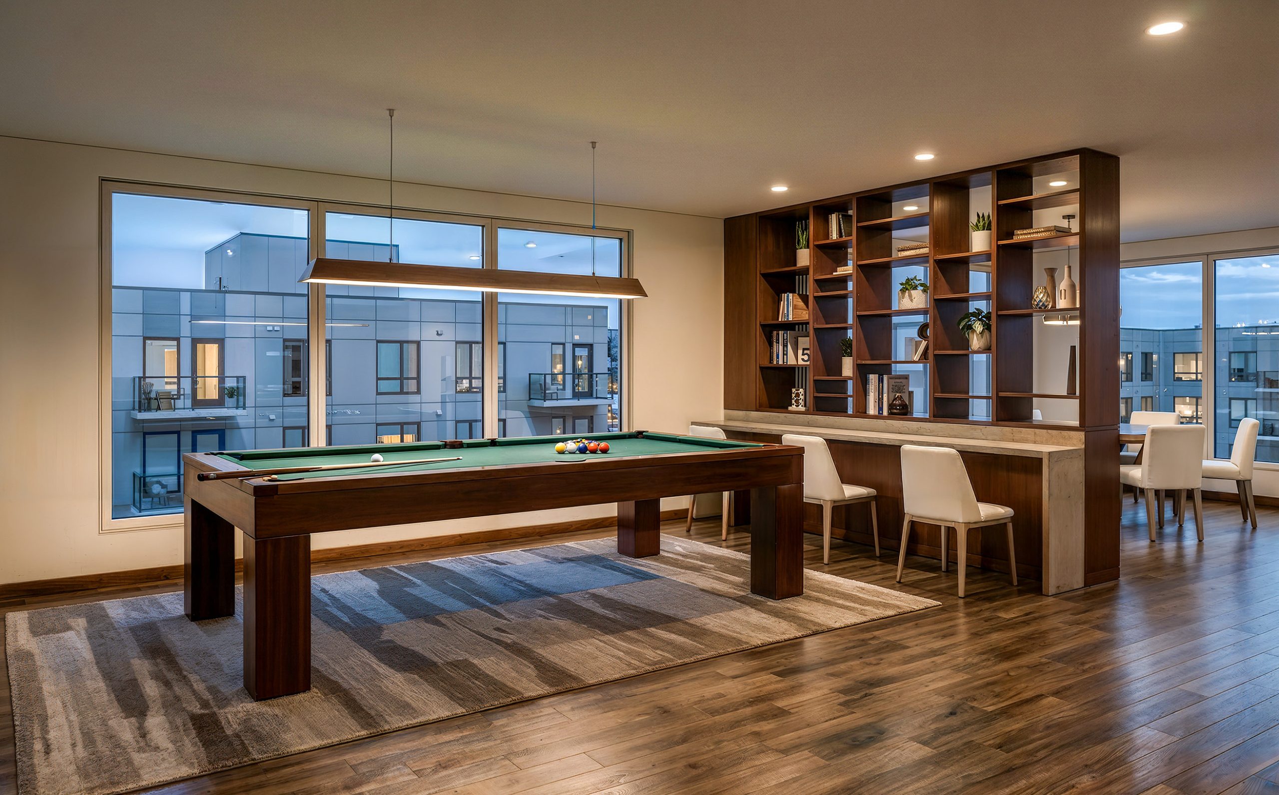 Hyde Square Bellevue game room and lounge interior rendering by Bogza.com. A contemporary resident amenity space featuring a green billiards pool table, warm wood flooring, a built-in room divider with decorative shelving, and bar seating looking out the window.