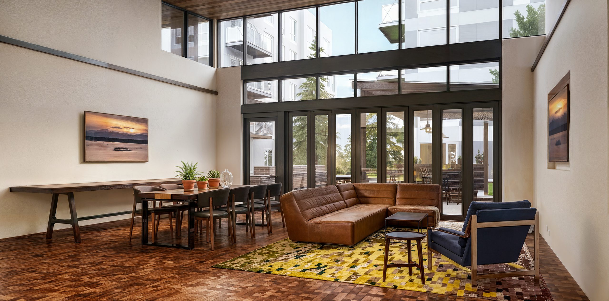 Hyde Square Bellevue resident lounge and clubhouse interior rendering by Bogza.com. A spacious indoor-outdoor amenity area featuring a large brown leather sectional sofa, a long dining table, and massive folding glass doors opening to a landscaped courtyard.