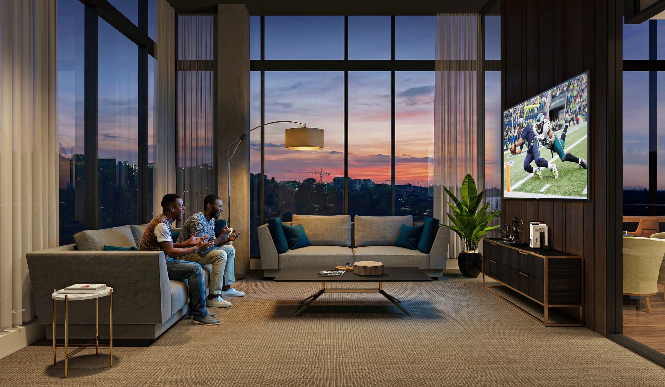 KODA Condominiums Seattle media lounge and screening room interior rendering by Bogza.com. A cozy, modern entertainment amenity space featuring residents relaxing on a comfortable grey sofa watching a football game on a large flat-screen TV, set against a dramatic sunset city view.