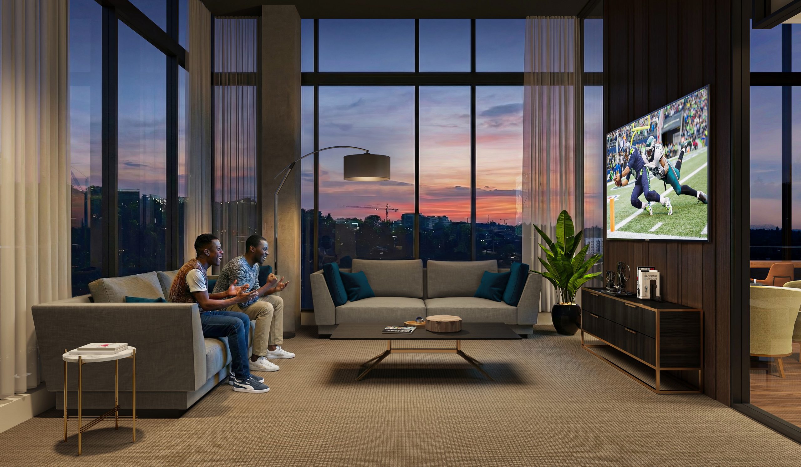 KODA Condominiums Seattle media lounge and screening room interior rendering by Bogza.com. A cozy, modern entertainment amenity space featuring residents relaxing on a comfortable grey sofa watching a football game on a large flat-screen TV, set against a dramatic sunset city view.