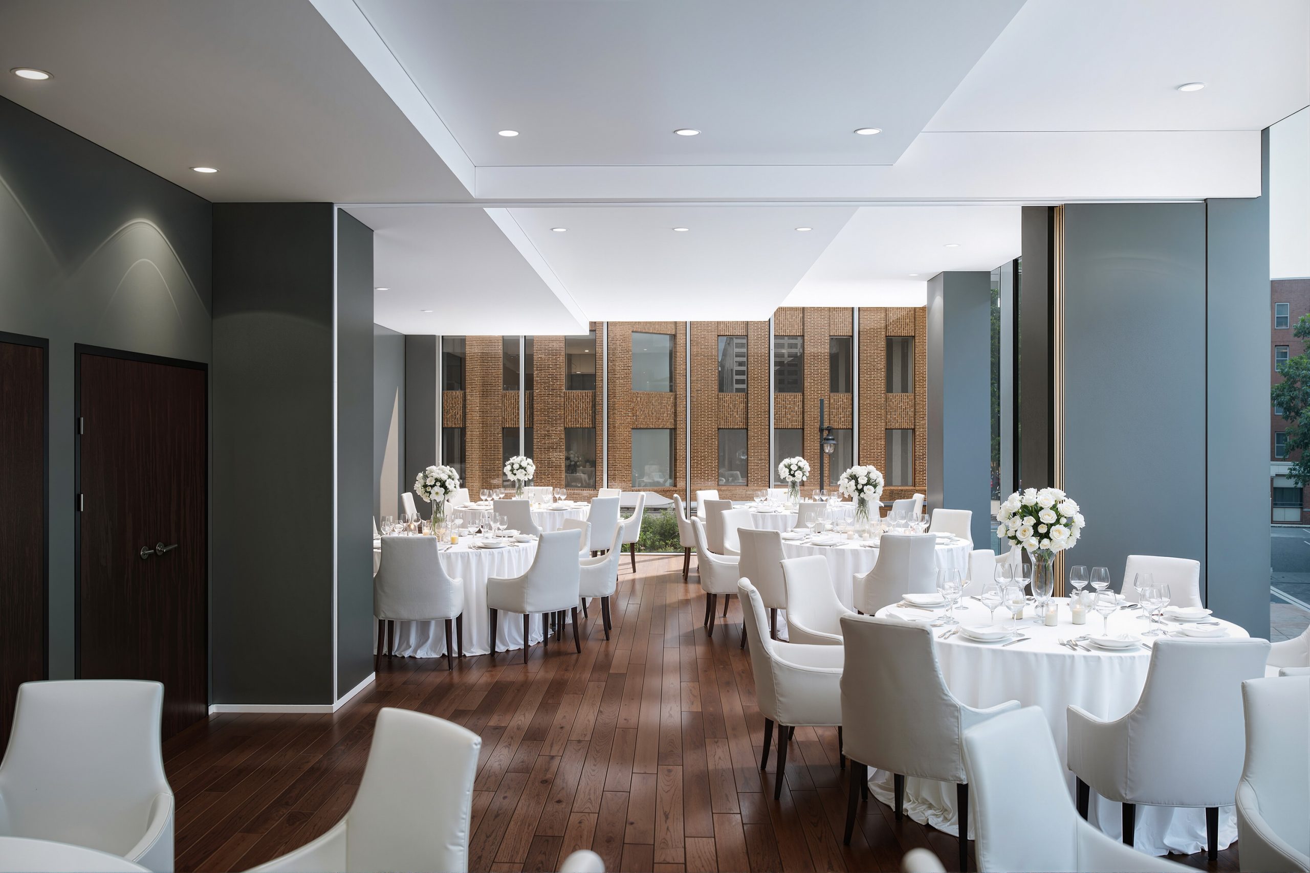 Thompson Seattle Hotel private event space and dining room interior rendering by Bogza.com. A sophisticated banquet room featuring round tables set with white linens and elegant floral centerpieces, warm hardwood flooring, modern seating, and large windows looking out onto the city.