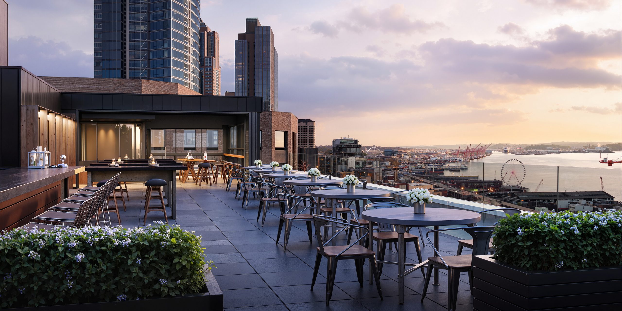 Thompson Seattle Hotel The Nest rooftop bar amenity rendering by Bogza.com. A stunning sunset view from the luxury hotel's outdoor lounge, featuring modern dining tables, bar seating, lush planters, and panoramic, unobstructed views of Elliott Bay and the Olympic Mountains.