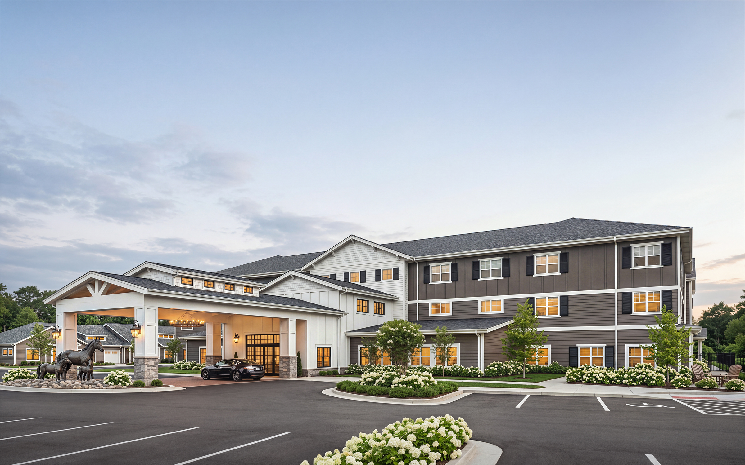 University Village senior living community main entrance architectural rendering by Bogza.com. A twilight street-level view of the luxury retirement campus, featuring classic exterior architecture with dark siding, a welcoming white porte-cochere, and a bronze horse sculpture.