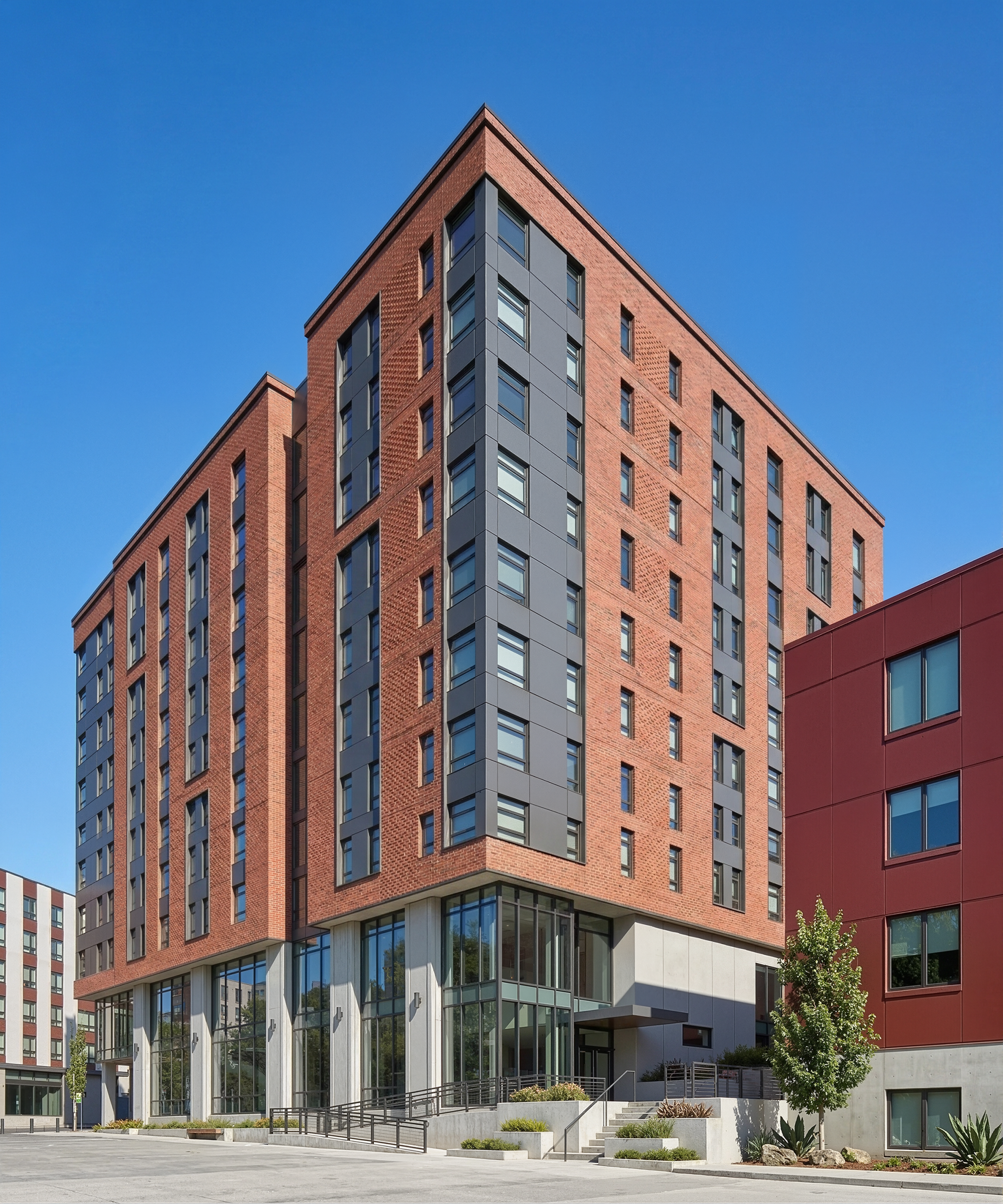Vi Hilbert Hall Seattle University architectural exterior rendering by Bogza.com. A bright daytime street-level corner view of the 10-story student residence building, featuring a modern facade with red brick and dark metal panels, large ground-floor windows, a concrete base, and an accessible entrance.