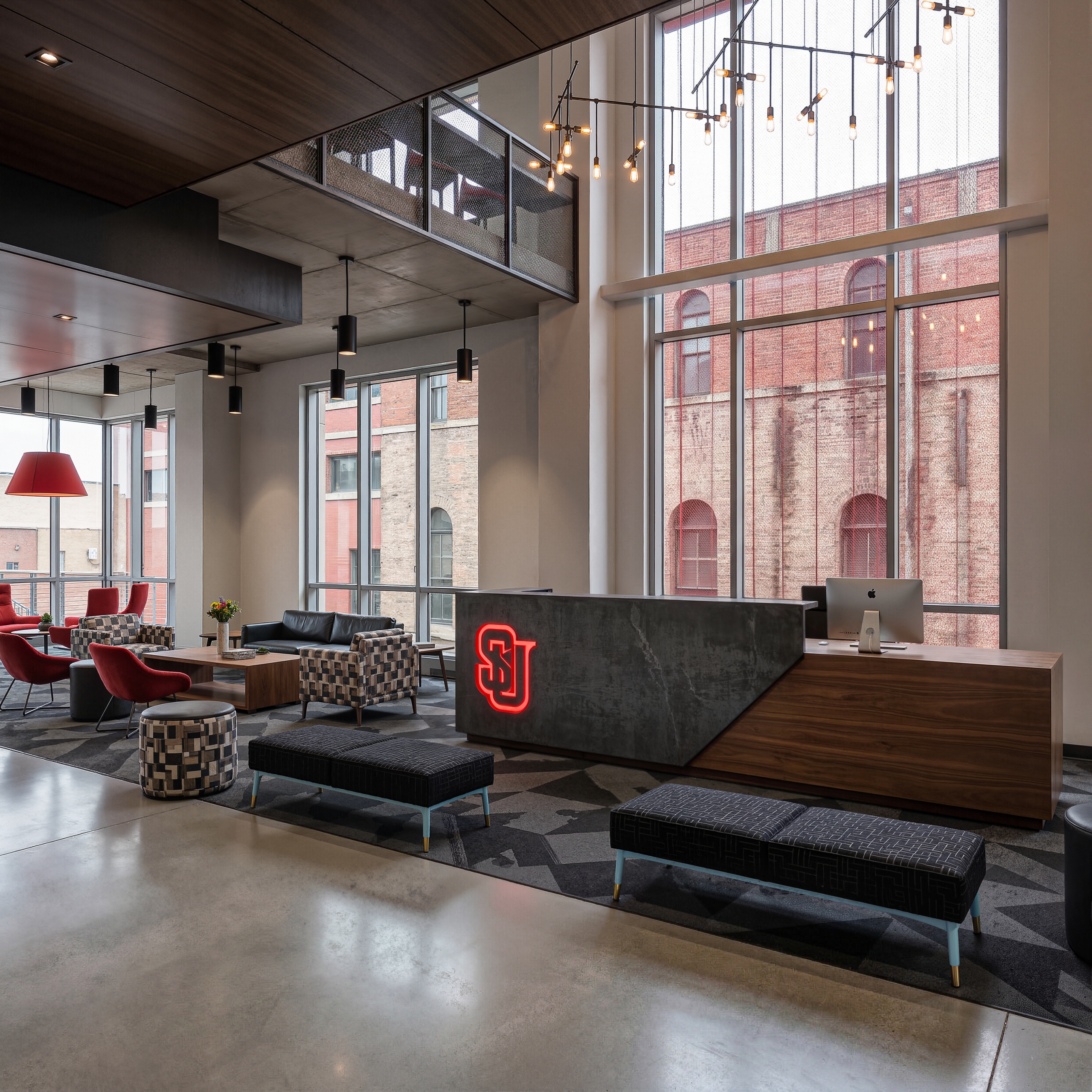 Vi Hilbert Hall Seattle University luxury lobby interior architectural rendering by Bogza.com. A bright, double-height reception area featuring a modern concierge desk with a glowing red SU logo, exposed concrete flooring, comfortable lounge seating, and expansive windows.