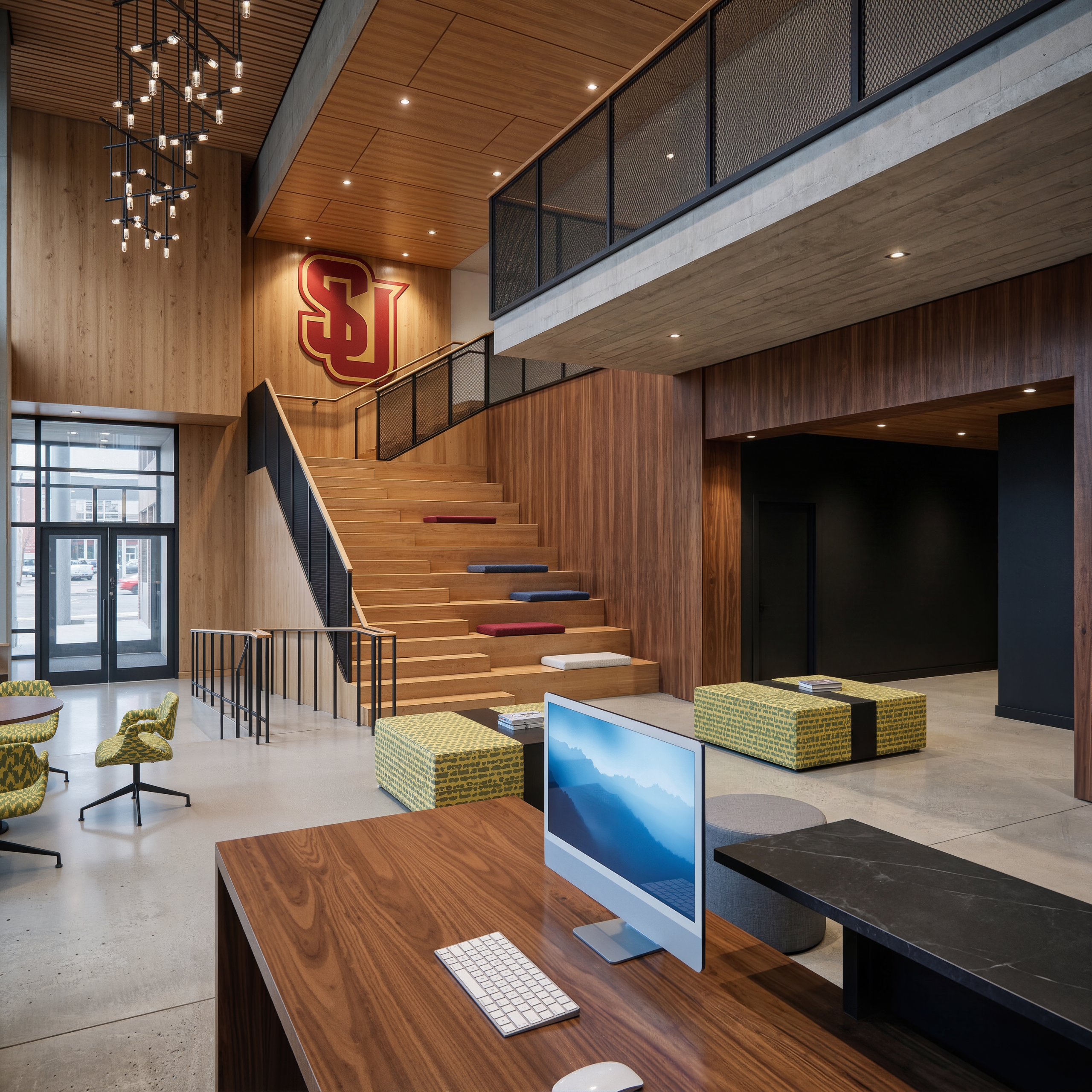 Vi Hilbert Hall Seattle University student lounge interior rendering by Bogza.com. A modern communal gathering space featuring a large wooden stadium-seating staircase with colorful cushions, warm wood paneling, a large Seattle U logo, and a computer workspace.